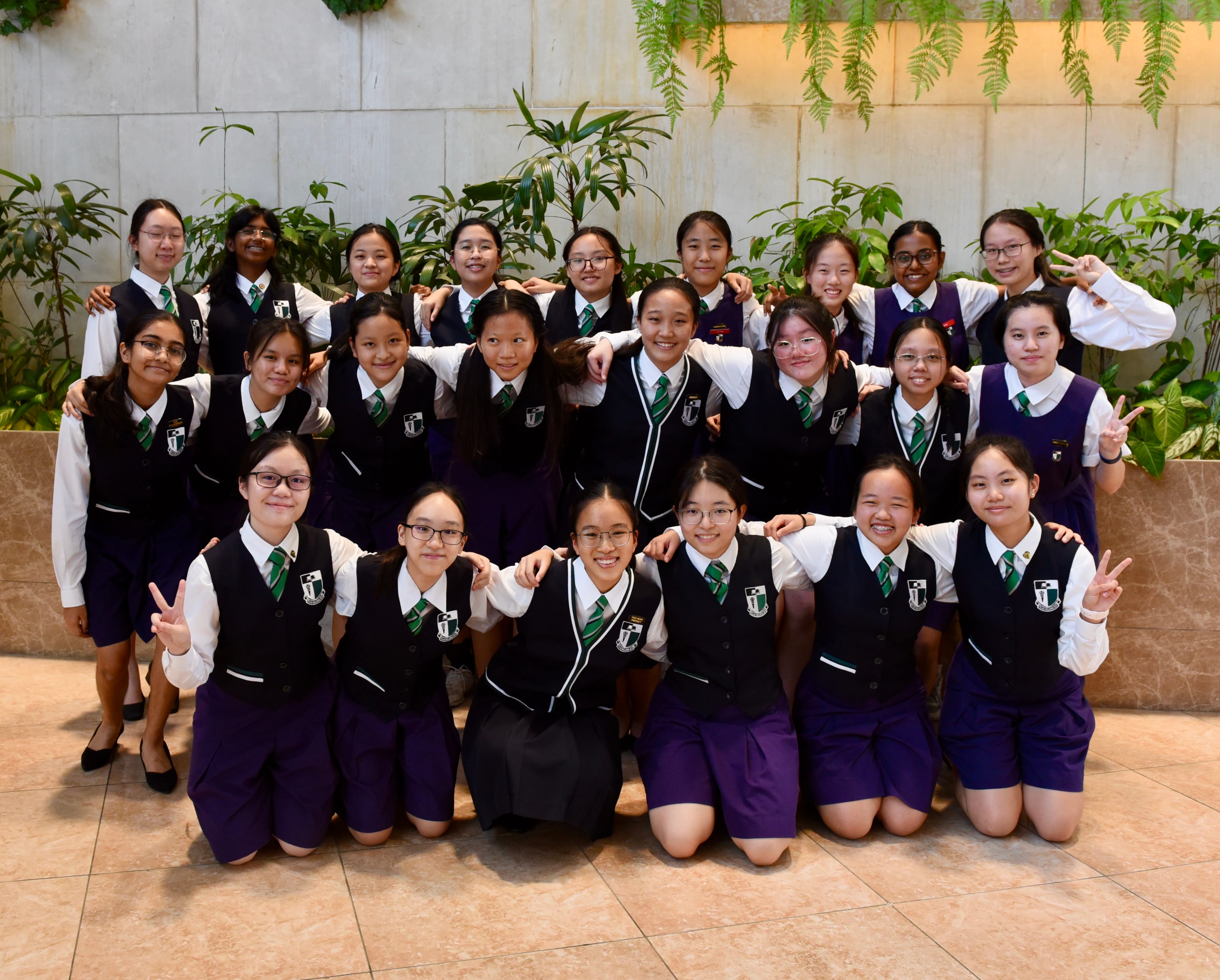 A group of about 20 schoolgirls in navy and white uniforms with green ties posing together indoors near plants.